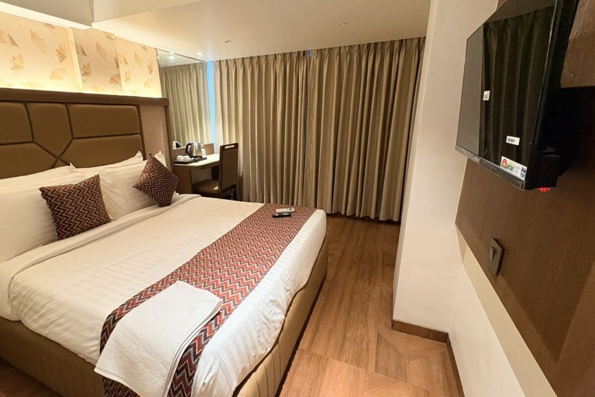 Habitación de hotel con una cama grande, un escritorio, una silla, un televisor y vistas al exterior a través de cortinas.
