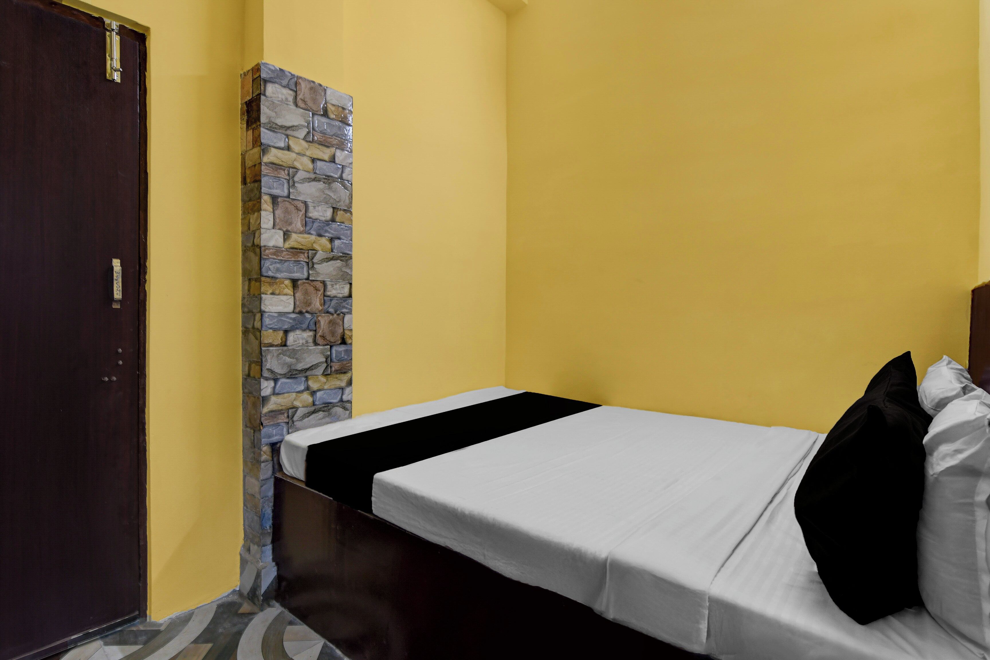 A single bed with a white mattress and black pillows, a stone accent wall, and a dark wooden door.