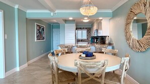 A dining area with a round table and wooden chairs, a chandelier, and a kitchen in the background. - 8th Floor Oasis! Beautiful Gulf front views! (Panama City Beach)