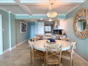 A dining area with a round table and wooden chairs, a chandelier, and a kitchen in the background. - 8th Floor Oasis! Beautiful Gulf front views! (Panama City Beach)