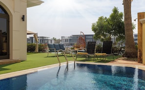 Une piscine entourée de chaises longues et d’un fauteuil suspendu, le tout bordé d’une pelouse bien entretenue et avec un bâtiment visible au loin.
