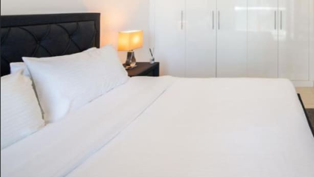 Uma cama bem arrumada com lençóis brancos, uma luminária de cabeceira e um guarda-roupa com duas portas.