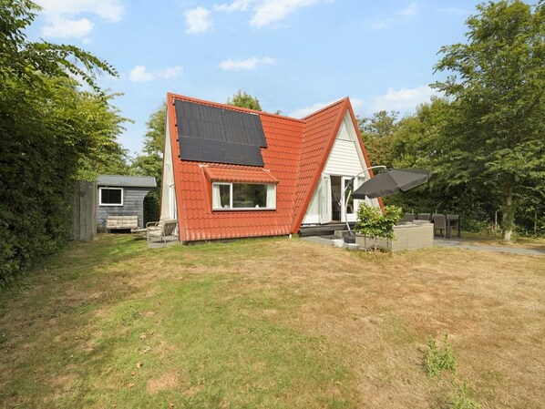 Ein Haus mit geneigtem Dach, Solaranlagen, einer kleinen Terrasse und einer Sitzgelegenheit im Freien.