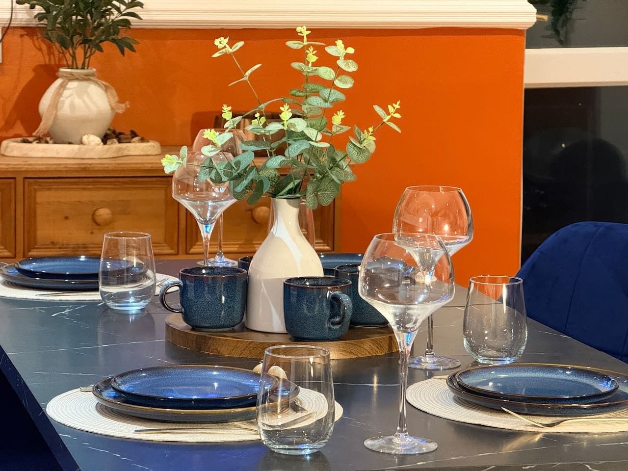 Une table à manger dressée avec des assiettes bleues, des verres transparents et un vase blanc contenant des plantes vertes.