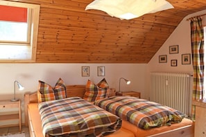 A bedroom with two beds, wooden ceiling beams, a window with blinds, a radiator, and a bedside table with a lamp. - Devil's tail||Lenzkirch (Lenzkirch)