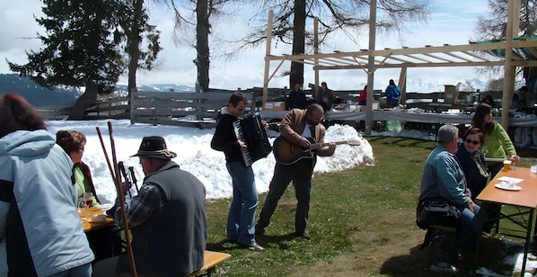An outdoor gathering with people sitting at tables, some playing musical instruments, and a wooden structure in the background.