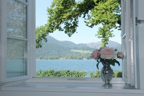 Ein Fenster mit Blick auf einen See und Berge, eine Vase mit Blumen auf dem Fensterbrett.