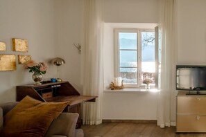 Ein Wohnzimmer mit Fernseher, Schreibtisch, Sofa und Fenster mit Blick ins Freie.