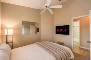 Ein Hotelzimmer mit Bett, Deckenventilator, an der Wand befestigtem Fernseher mit 'NETFLIX'-Anzeige und Nachttisch, sowie einer Tür zu einem weiteren Raum.