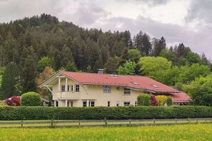 Una casa de dos pisos con techo de tejas rojas, rodeada por un seto cuidado y un campo de flores amarillas.