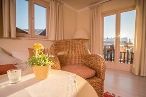 A room with a wicker chair, a table with a glass, and a view of buildings through the window. - in the Riesch guesthouse | holiday apartment (Garmisch-Partenkirchen)