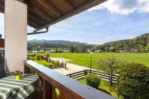 A balcony with a view of a green landscape, a wooden railing, and a table with a checkered tablecloth. - Melf "Summer linden" (Walchensee)