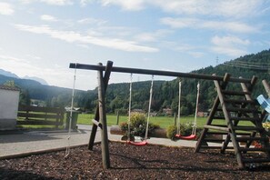 A playground with swings and a slide, surrounded by a fence and greenery. - Melf "Summer linden" (Walchensee)