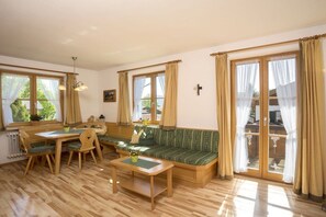 A room with a wooden dining table, chairs, a bench, and a sofa. There are windows with curtains, a radiator, and a cross on the wall. - Melf "Summer linden" (Walchensee)