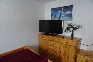 A bedroom with a wooden dresser, a television on top, a bed with a red blanket, and a wall-mounted painting of a tropical scene. - Holiday apartment with 2 bedrooms (Tambach-Dietharz)