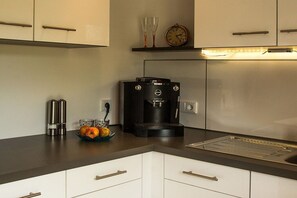 Eine moderne Küche mit weißen Schränken, einer Kaffeemaschine, einer Obstschale und einer Uhr an der Wand.
