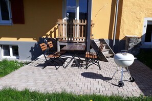 En udendørs patio med et træbord og bænke, en grill og en gul bygning i baggrunden.