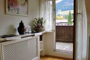 Ein Zimmer mit Heizkörper, einer Pflanze auf einem Fensterbrett, einem Laptop und Blick auf eine Holzterrasse und Berge.