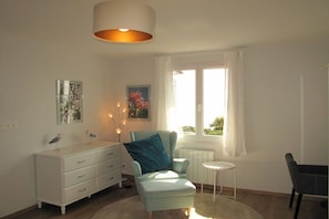 Ein Zimmer mit einer weißen Kommode, einem türkisnen Sessel, einem kleinen runden Tisch und einem Fenster mit Vorhängen.