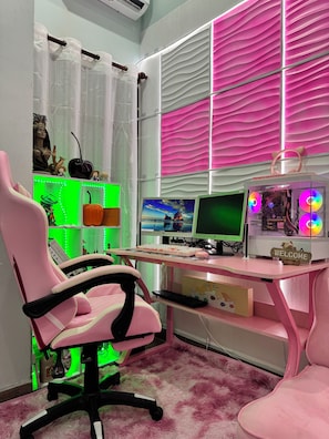 Una habitación con una silla gamer rosa, un escritorio con equipo de computadora y una alfombra suave color rosa.