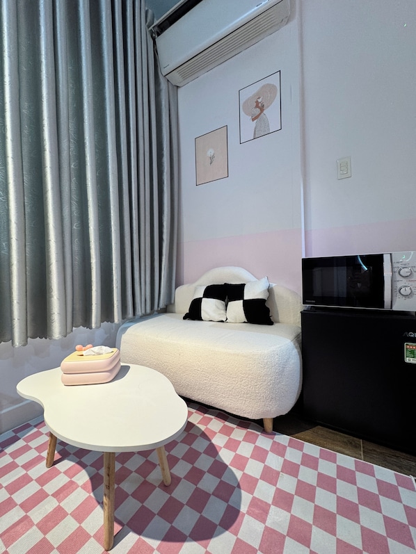 A room with a checkered floor, a white sofa with black and white cushions, a small table with a pink object, a black TV stand, and a window with grey curtains.