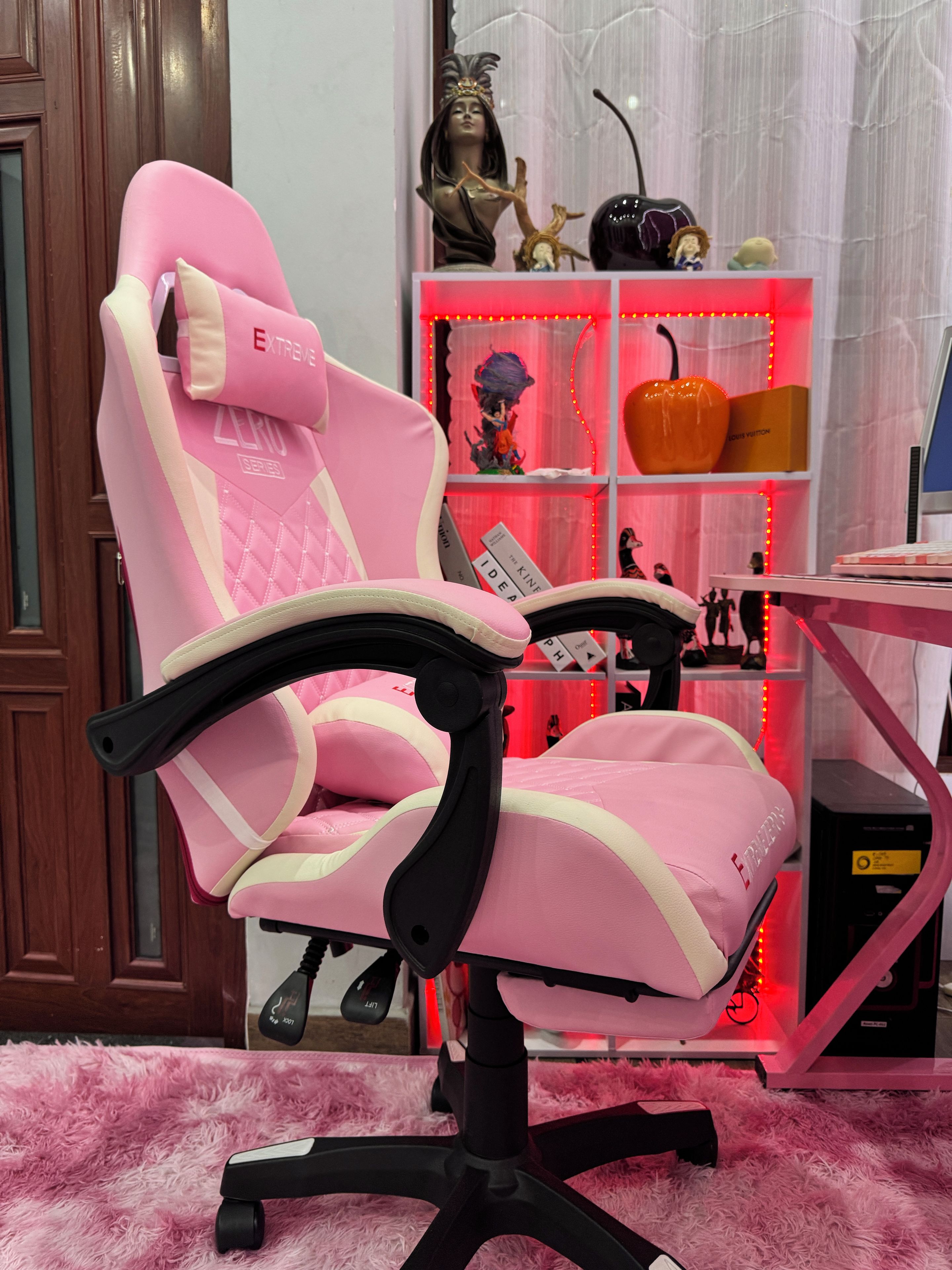 Una silla gamer rosa con reposabrazos y respaldo alto, ubicada frente a un escritorio con un monitor de computadora.