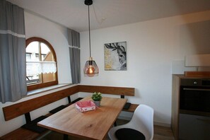 A dining area with a wooden table, benches, a hanging light fixture, a window with curtains, and a kitchen area with a stove. - Alpenflair Holiday Apartment 232 (Oberstdorf)