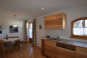 A kitchen with wooden cabinets, a dining table with chairs, a window with curtains, and a hanging light fixture. - Alpenflair holiday apartment 241 (Oberstdorf)
