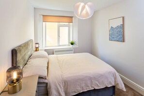 Ein Schlafzimmer mit Bett, Nachttisch, Lampe, Fenster mit Jalousien, Heizkörper und einem gerahmten Bild an der Wand.