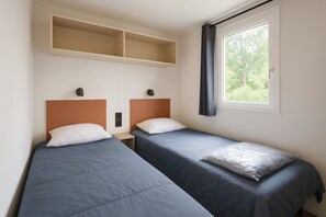 A hotel room with two single beds, a window with curtains, and a shelf above the beds. - Mobile Home '6 Pers Domaine La Yole' with Shared Pool, Private Terrace and Air Conditioning (Vendres)