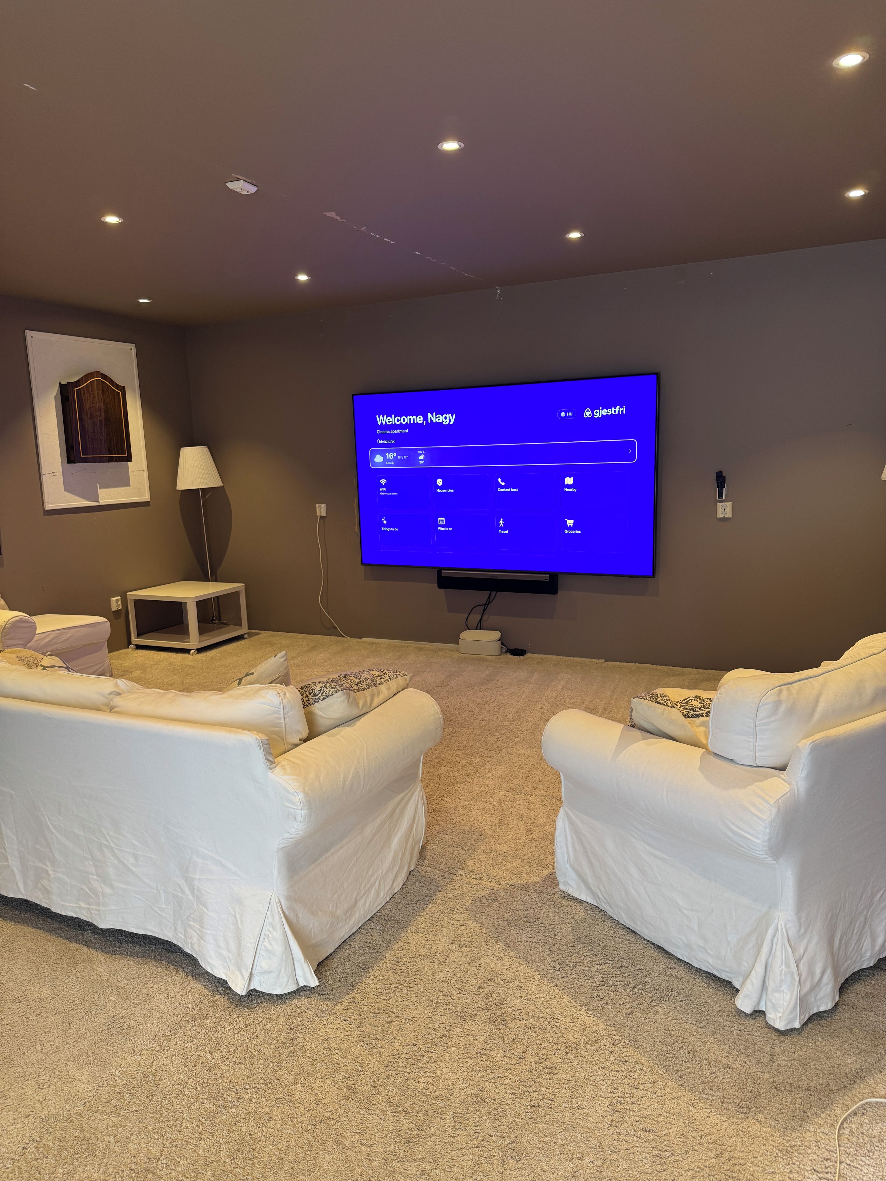 Sala de estar com televisão de tela plana exibindo uma mensagem de boas-vindas, dois sofás brancos, um abajur e uma pequena mesa.