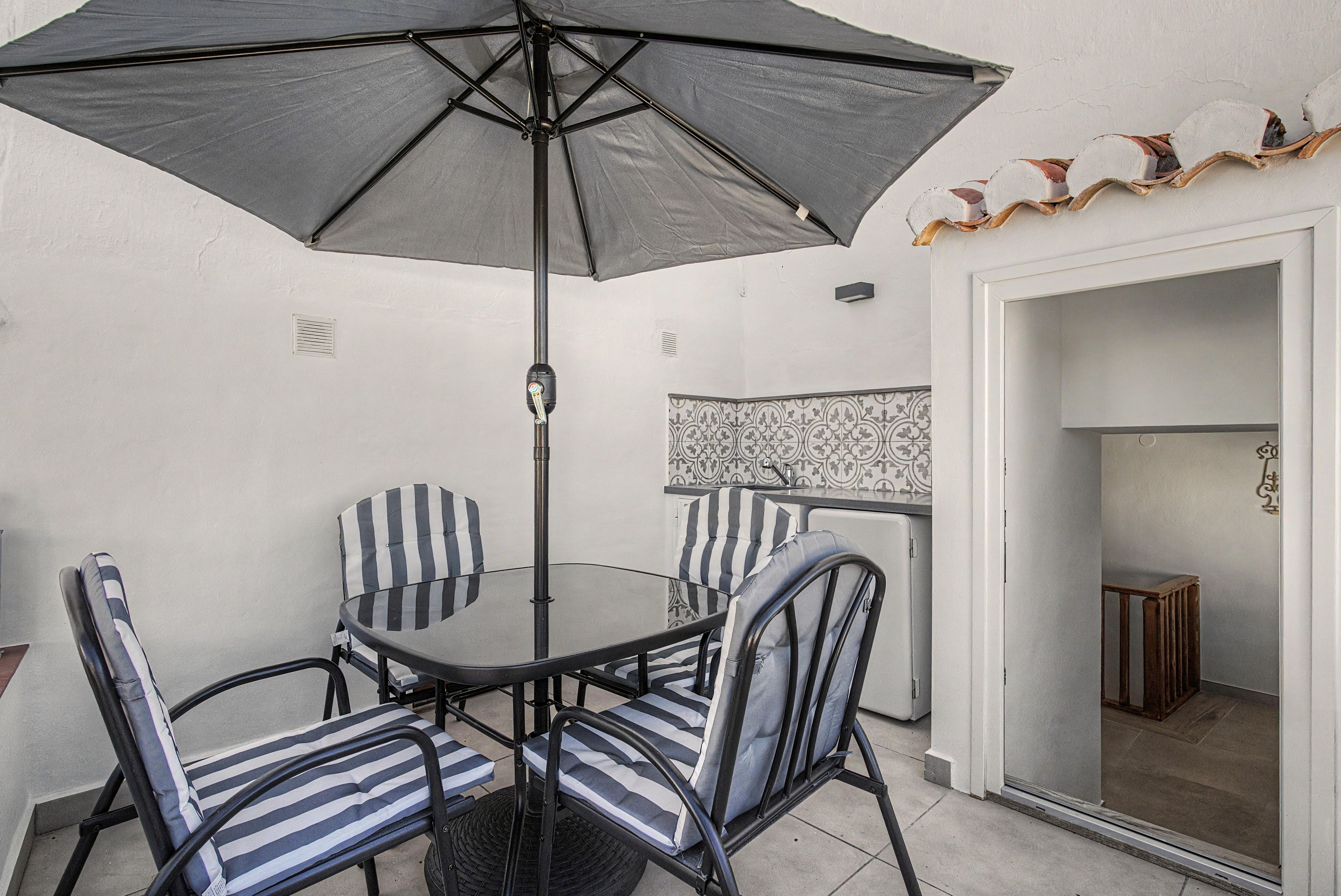 A dining area with a round table, four striped chairs, a large umbrella, and a patterned backsplash.