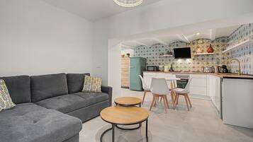 A modern living room with a grey sofa, a round wooden table, and a kitchen area with a TV, microwave, and patterned backsplash.