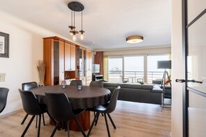 A modern dining area with a round table and chairs, a wooden cabinet, and a view of the sea through large windows. - Apartment in Zeedijk With Stunning Sea Views (Blankenberge)