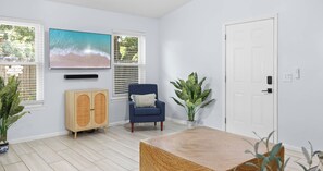 A room with a large window, a blue armchair, a wooden cabinet, and a television on the wall. - Pinetree Palace - 4BR Cozy Home (Panama City Beach)