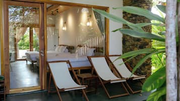 Een slaapkamer met een bed, twee houten stoelen, een stenen muur en een glazen deur die leidt naar een balkon.