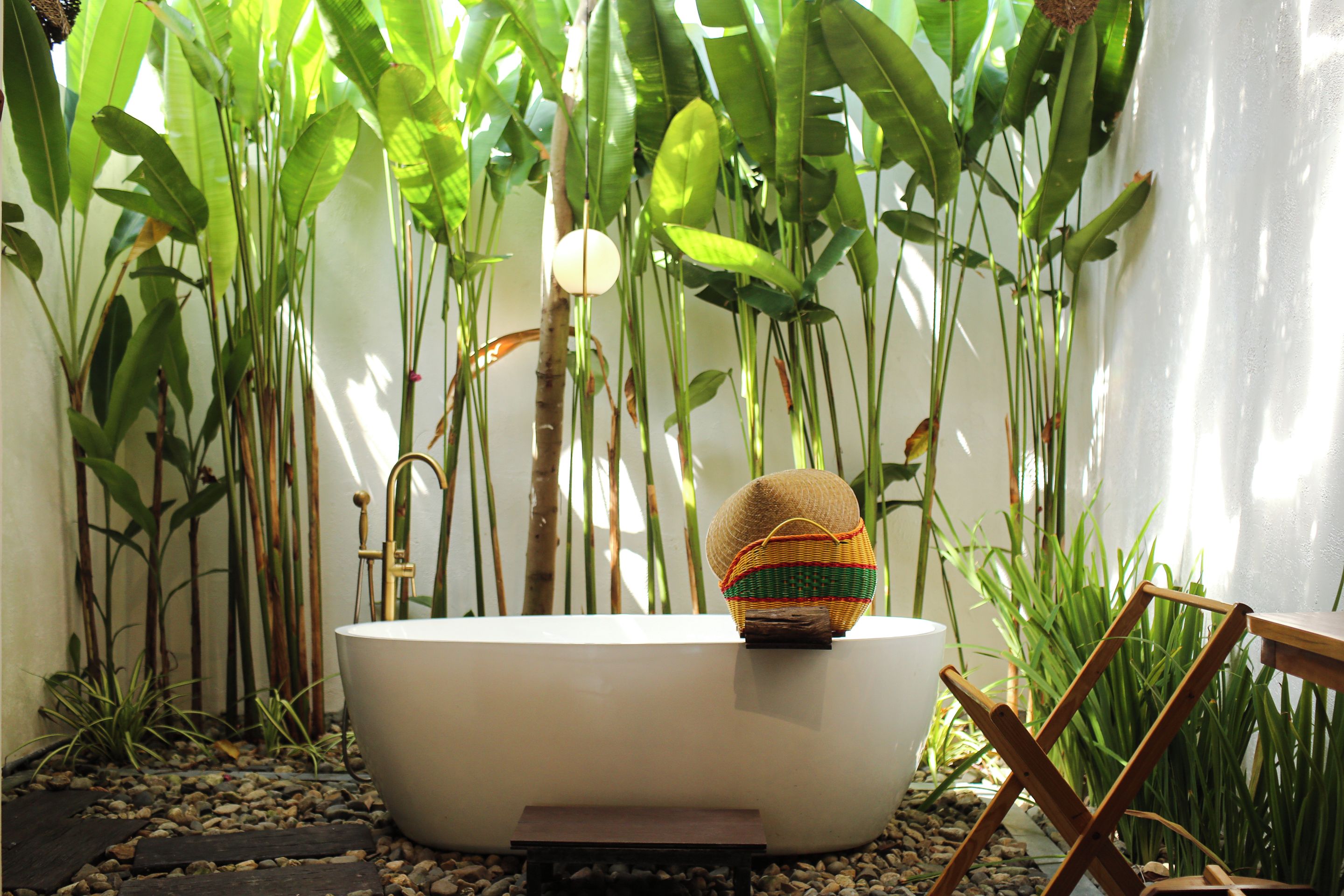 Una bañera con una cesta tejida encima, rodeada de plantas verdes altas y una silla de madera.