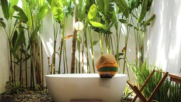 Una bañera con una cesta tejida encima, rodeada de plantas verdes altas y una silla de madera.