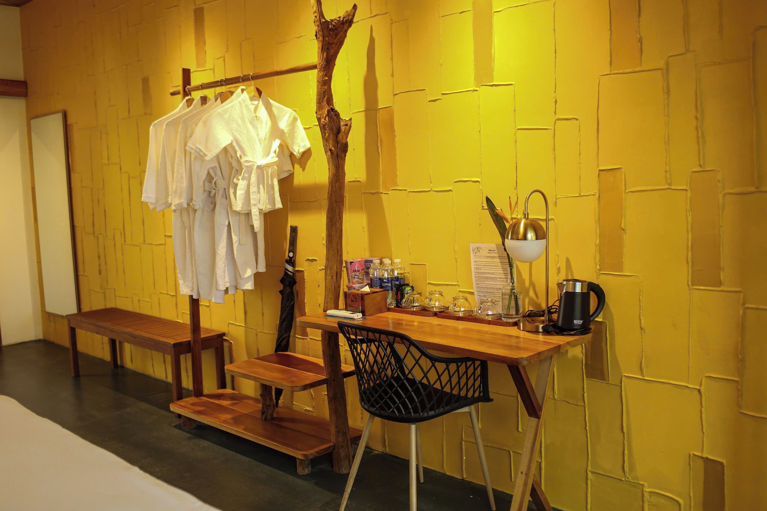 A room with a wooden bench, a table with a chair, a coat rack with clothes, a coffee maker, and a wall with a textured pattern.
