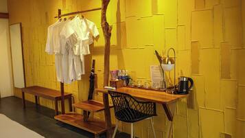 A room with a wooden bench, a table with a chair, a coat rack with clothes, a coffee maker, and a wall with a textured pattern.