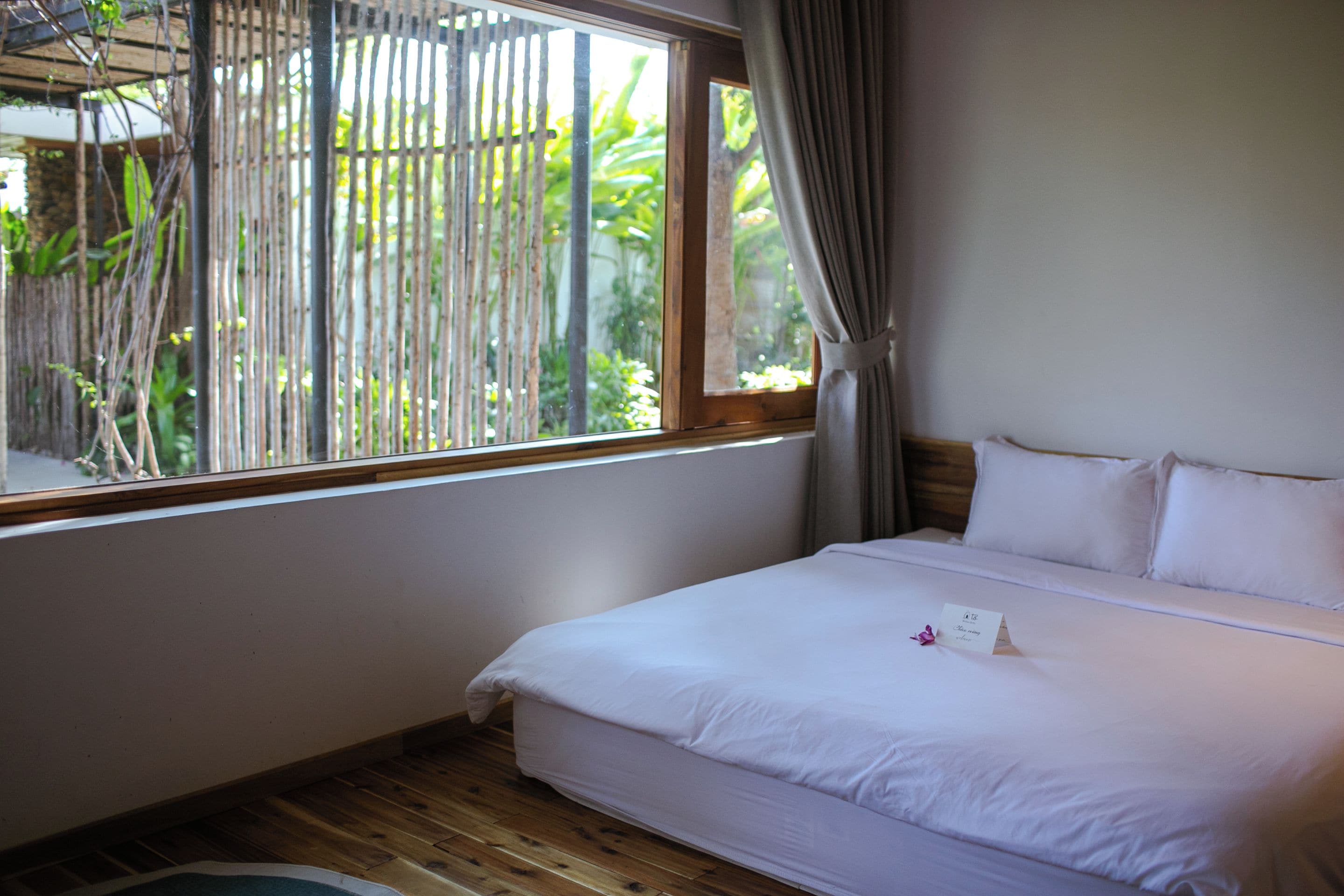 Quarto de hotel com uma cama, janela com vista para área verde e piso de madeira.