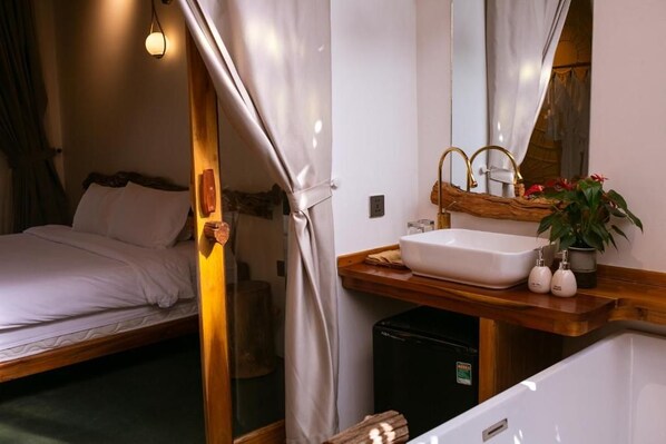 A hotel room with a bed, a wooden bedside table, a sink, and a mirror.