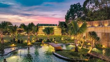 A serene outdoor pool area with a wooden deck, surrounded by palm trees and bamboo fencing, under a twilight sky.