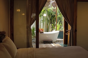 A bedroom with a view of a bathroom featuring a bathtub, surrounded by greenery.