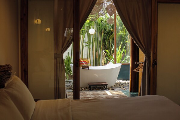A bedroom with a view of a bathroom featuring a bathtub, surrounded by greenery.