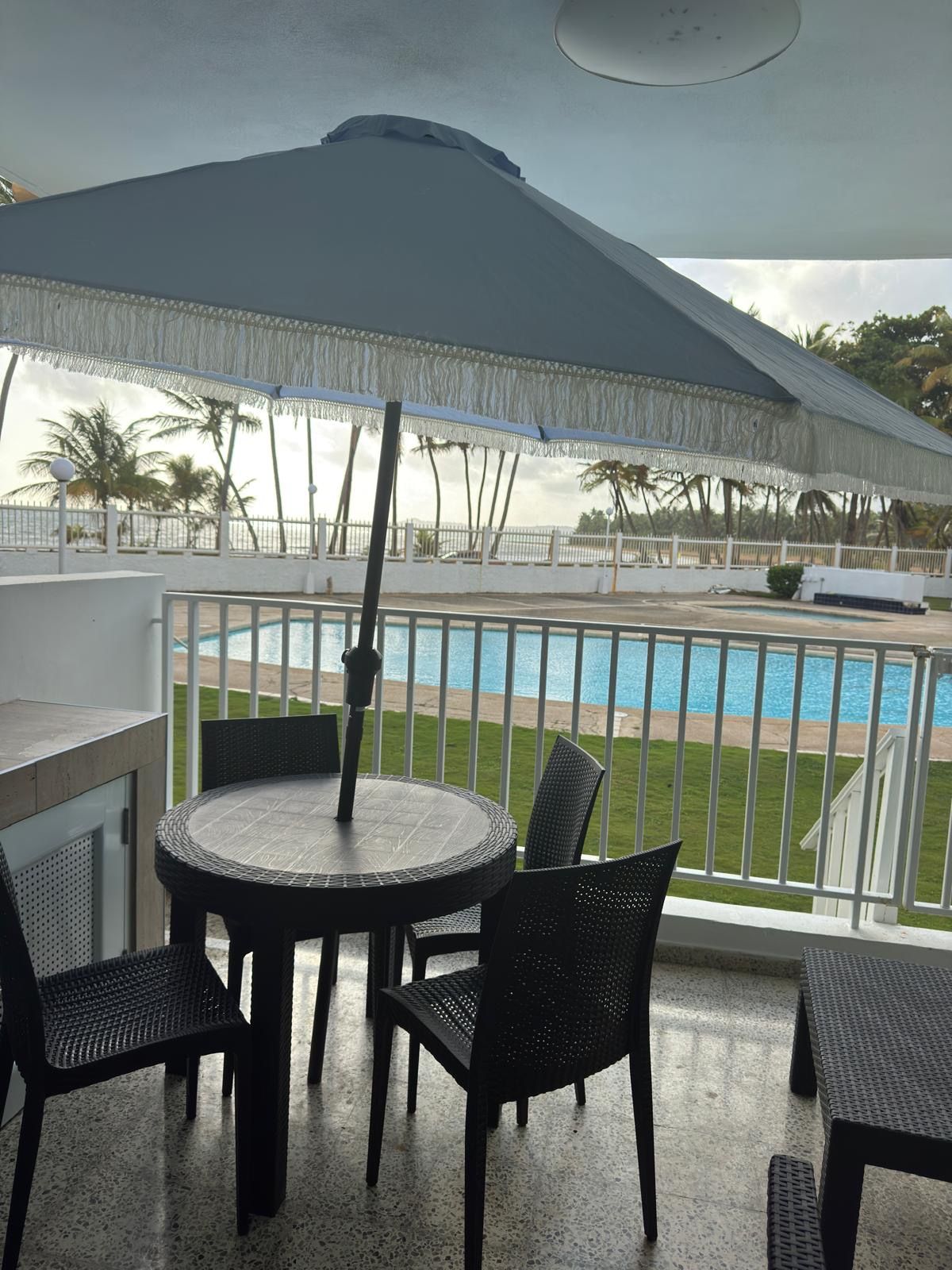 A covered outdoor seating area with a table and chairs, overlooking a pool and beach.