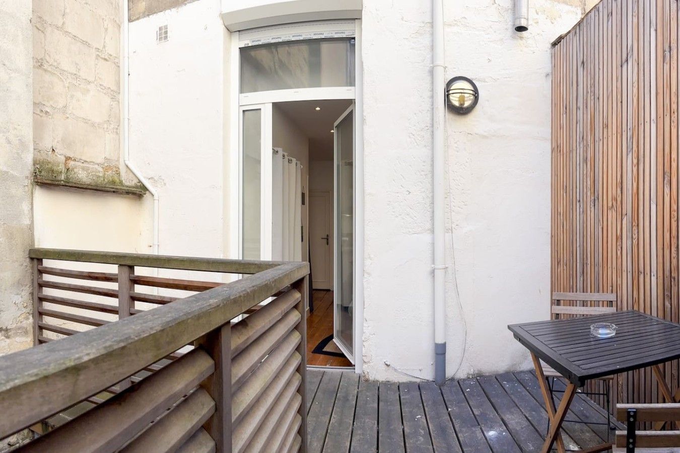 Une terrasse en bois avec une table et des chaises, un mur blanc avec une clôture en bois et une porte donnant sur un espace intérieur.