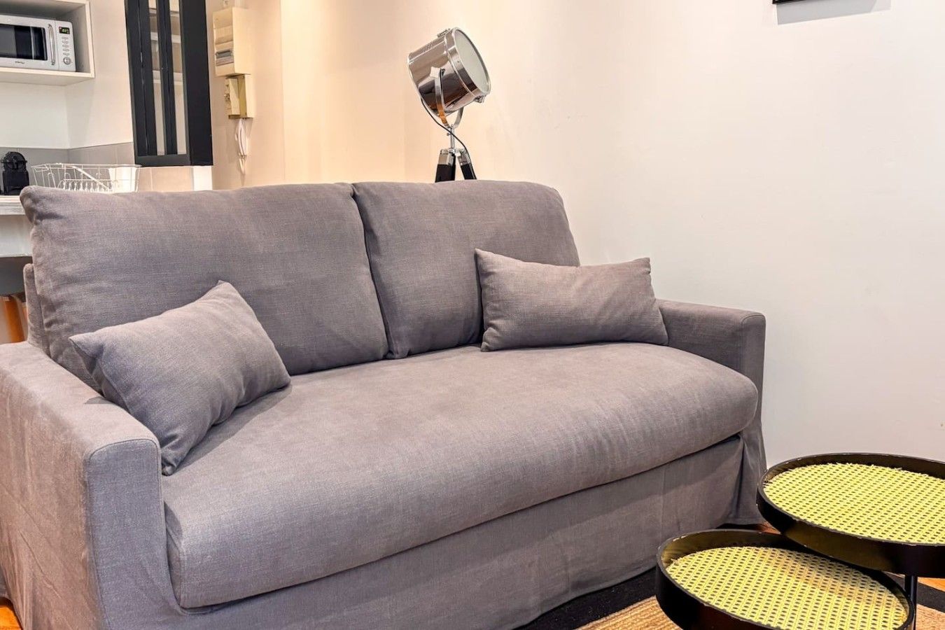 Un canapé gris accompagné d’un coussin assorti, une petite table ronde avec un plateau en fibres tressées et un lampadaire dans une pièce aux murs blancs, avec un coin cuisine en arrière-plan.