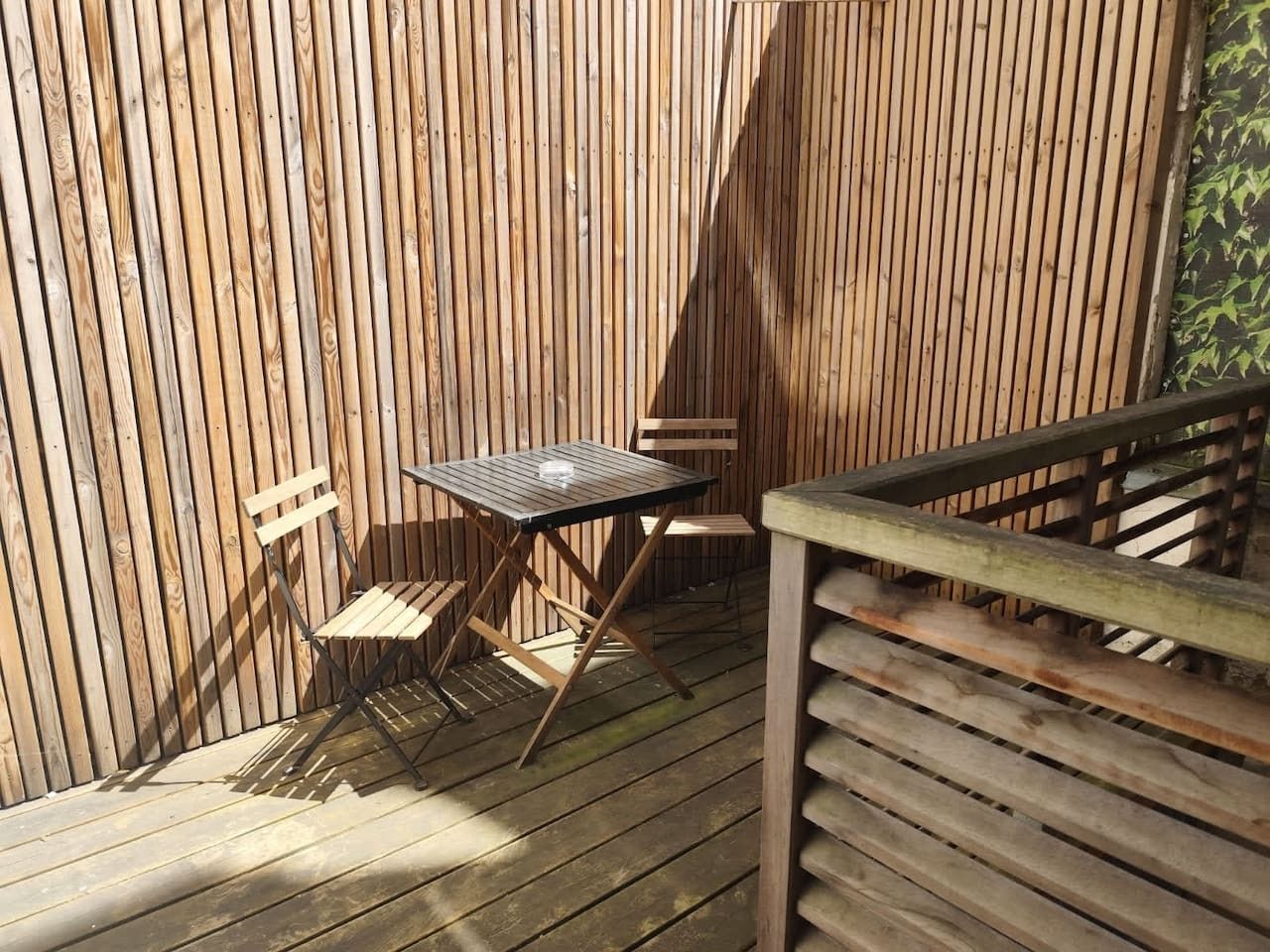 Une terrasse en bois avec une table et des chaises, une rambarde en bois et un mur composé de panneaux de bois verticaux.