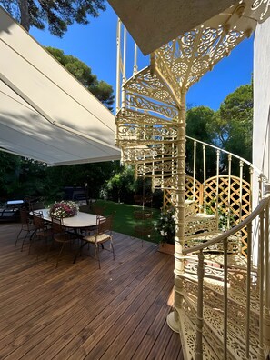 A wooden deck with a round table and chairs, surrounded by greenery and a white canopy.
